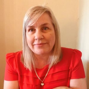 A person with light colored, shoulder-length hair wearing a bright red top with pleated detailing and a gold heart-shaped pendant necklace, seated indoors against a neutral background.